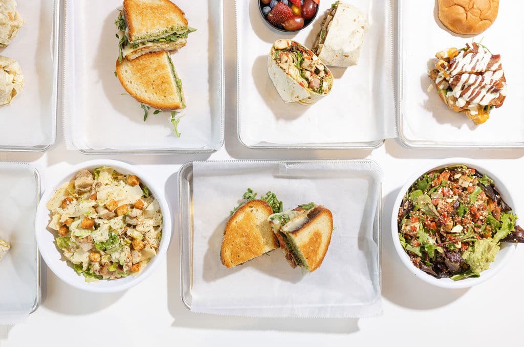 Overhead spread of Comfort Fuel grilled sandwiches, a chicken wrap, a loaded sandwich, Caesar salad and a quinoa bowl